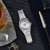 A silver-toned Lorier Astra wristwatch styled in a flat-lay arrangement on a dark textured surface with a pen, sunglasses, cufflinks, and a drink nearby.