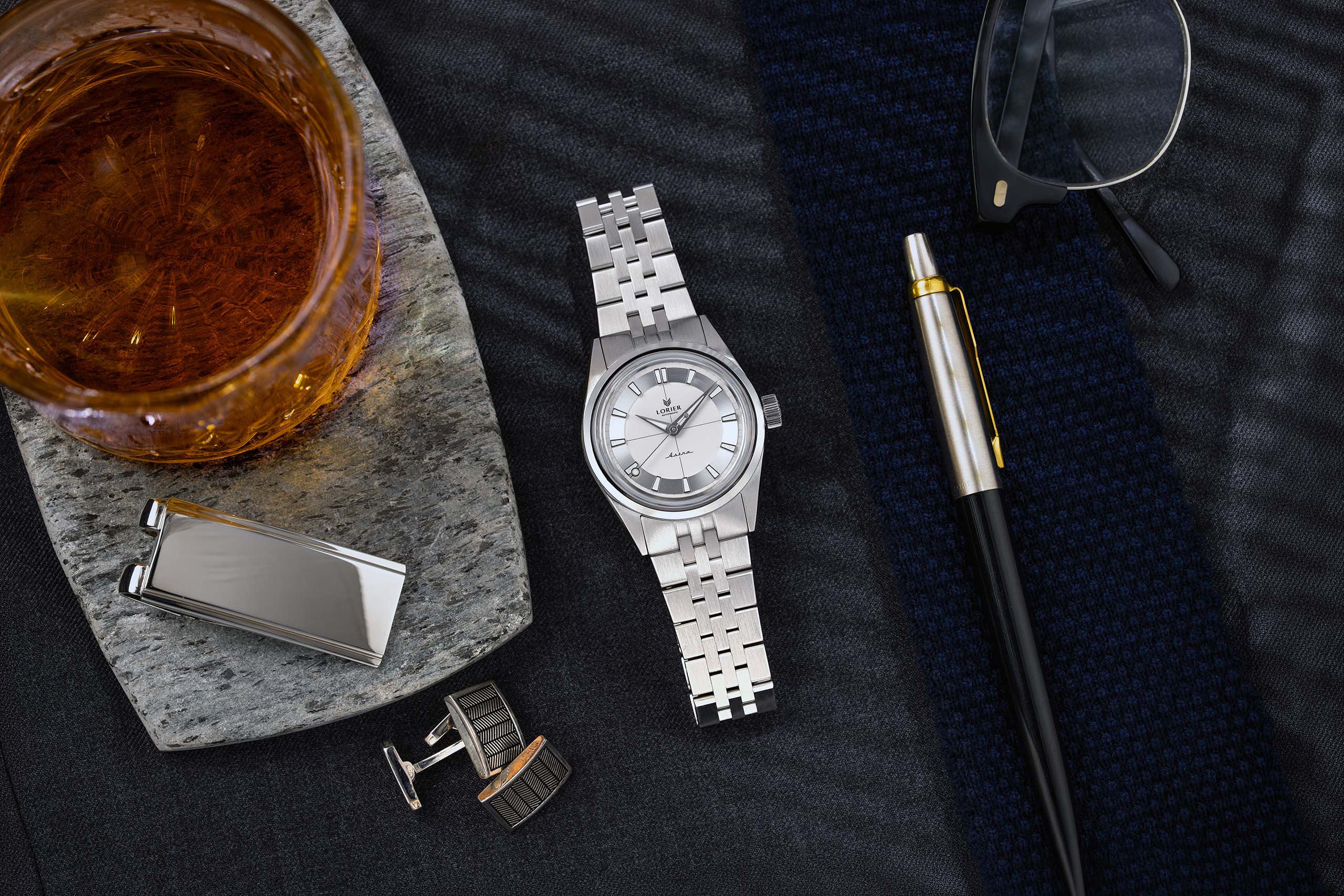A silver-toned Lorier Astra wristwatch styled in a flat-lay arrangement on a dark textured surface with a pen, sunglasses, cufflinks, and a drink nearby.