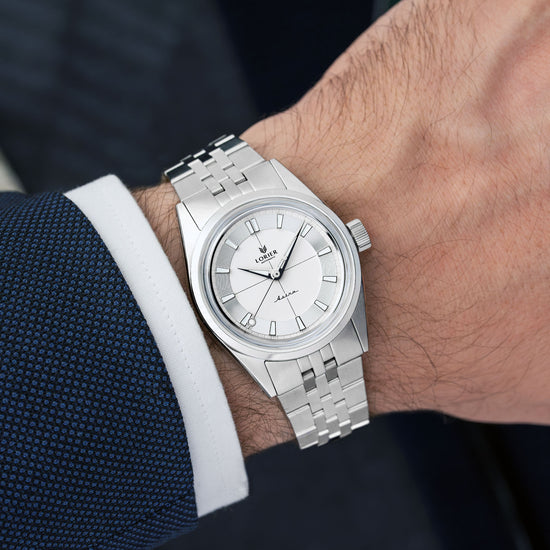 A Lorier Astra wristwatch with a white dial and five-link bracelet, shown worn on a person's wrist with a dark blue textured suit jacket and white dress shirt cuff visible.