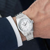 A Lorier Astra wristwatch with a white dial and five-link bracelet, shown worn on a person's wrist with a dark blue textured suit jacket and white dress shirt cuff visible.