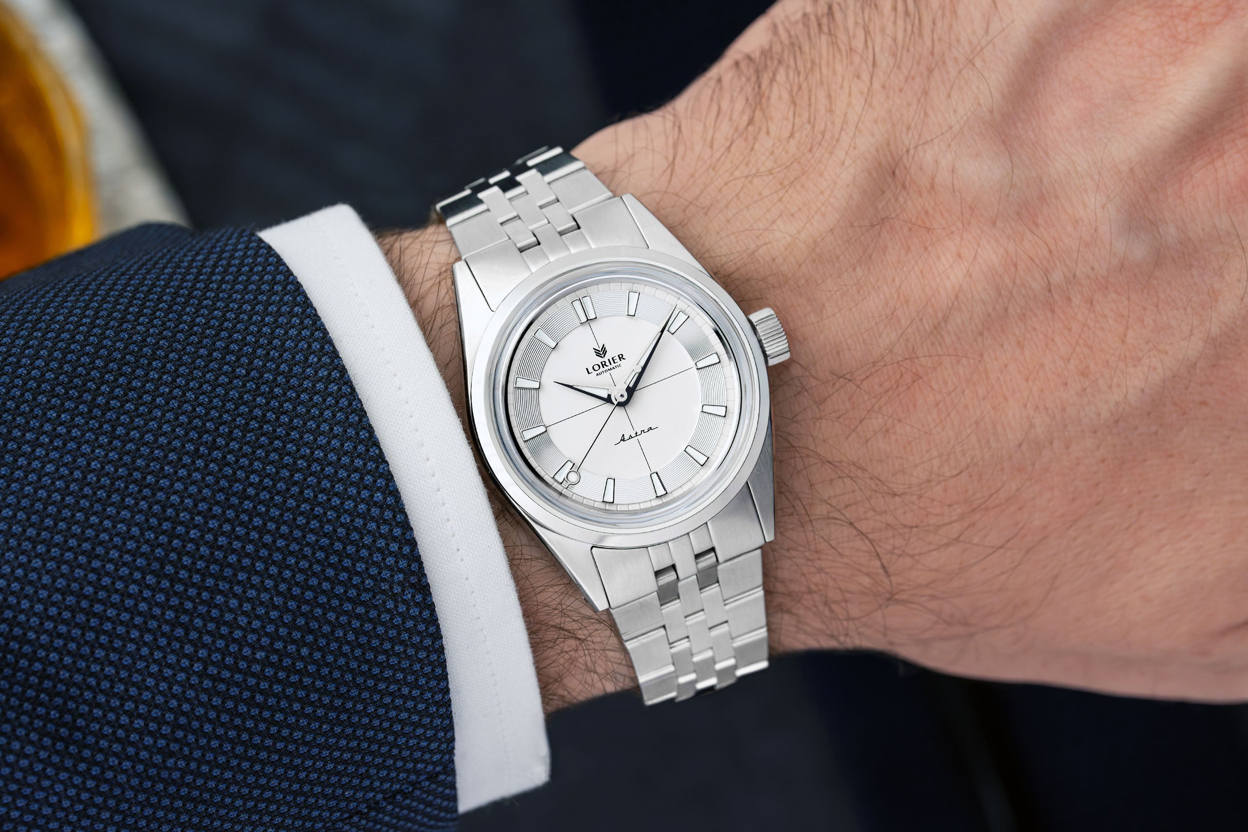 A Lorier Astra wristwatch with a white dial and five-link bracelet, shown worn on a person's wrist with a dark blue textured suit jacket and white dress shirt cuff visible.
