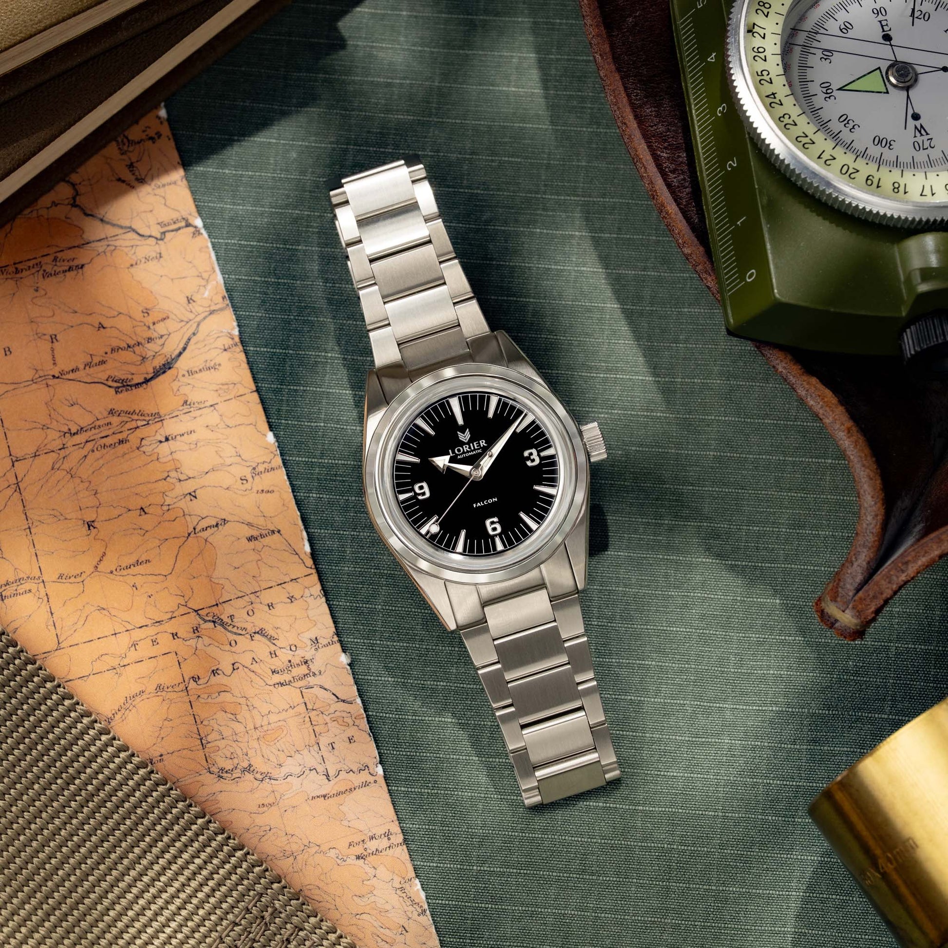 Lorier Falcon field watch flatlay on green canvas surface with vintage topographic map, military-issue compass in olive case, brass telescope, and leather field notebook.