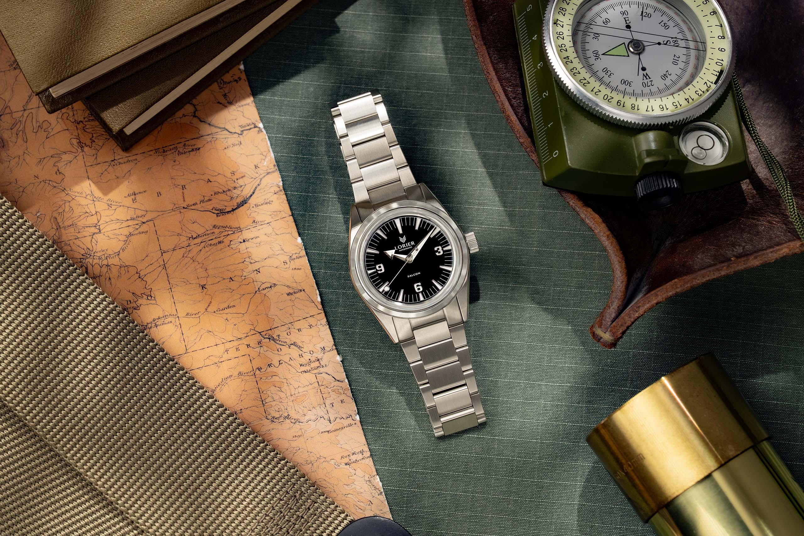 Lorier Falcon field watch flatlay on green canvas surface with vintage topographic map, military-issue compass in olive case, brass telescope, and leather field notebook.