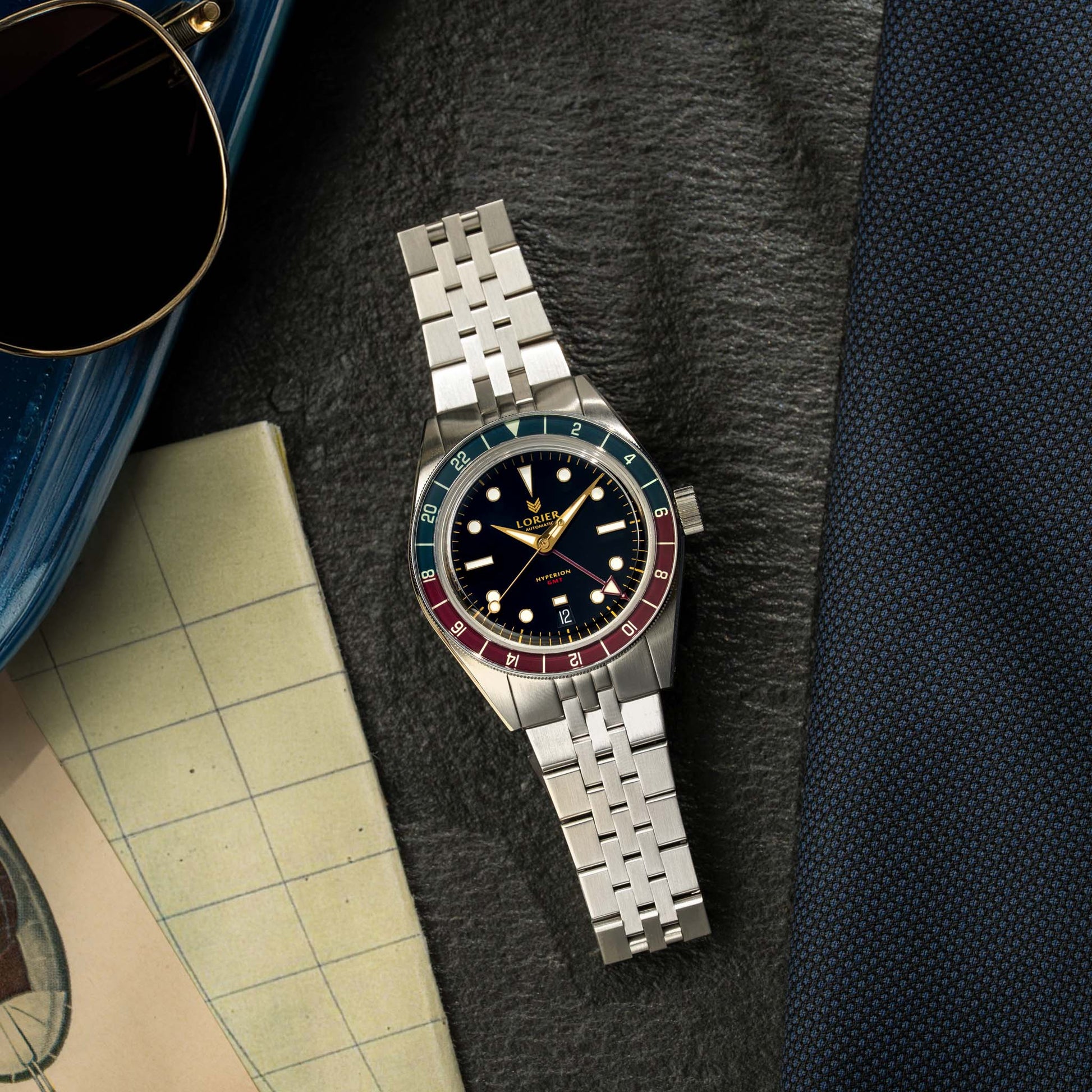 A Lorier Hyperion wristwatch with a black dial and a red-and-blue bezel sits on a dark textured surface, styled with aviator sunglasses, a blue case, a vintage map, and a navy suit jacket.