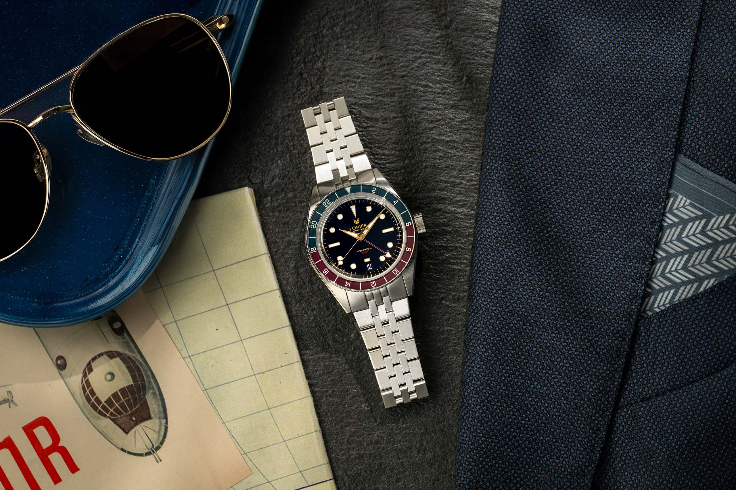 A Lorier Hyperion wristwatch with a black dial and a red-and-blue bezel sits on a dark textured surface, styled with aviator sunglasses, a blue case, a vintage map, and a navy suit jacket.
