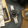 Lorier Neptune dive watch flatlay on grey leather surface with vintage nautical depth chart, U.S. Divers consumer catalog, and open antique brass surveying compass on aged craft paper. 