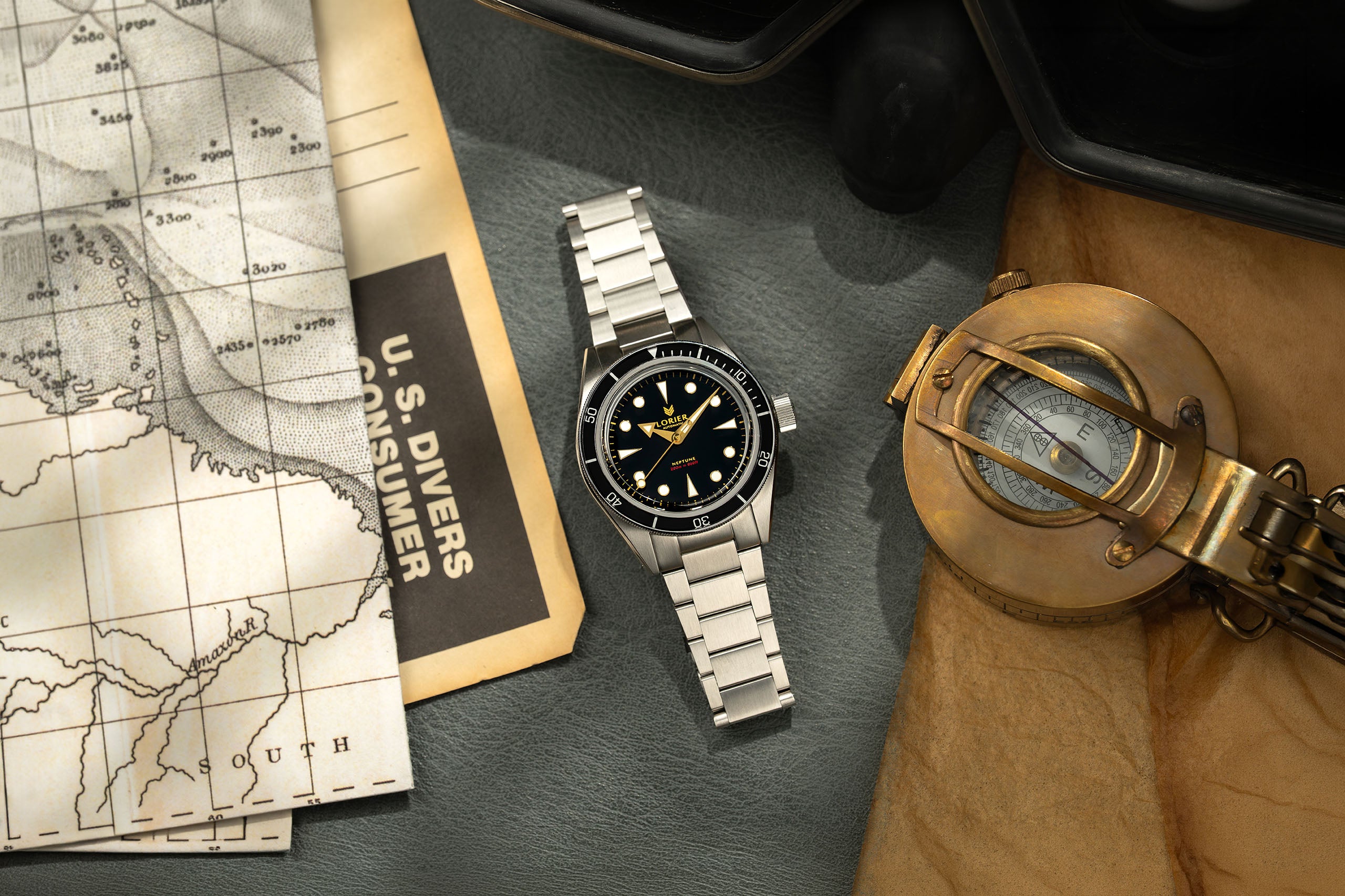 Lorier Neptune dive watch flatlay on grey leather surface with vintage nautical depth chart, U.S. Divers consumer catalog, and open antique brass surveying compass on aged craft paper. 